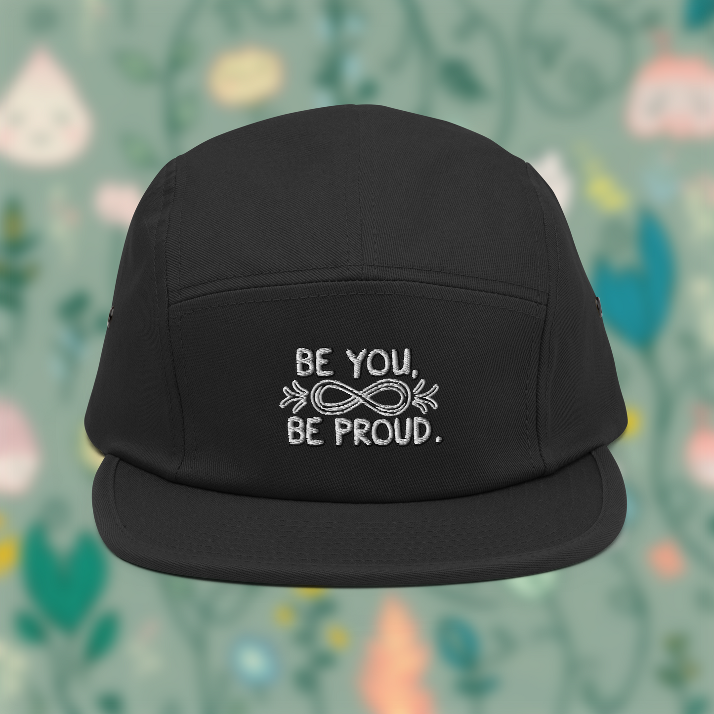 Black cap with embroidered text 'Be You, Be Proud' and an infinity symbol with arrows, set against a blurred background of pastel colours.