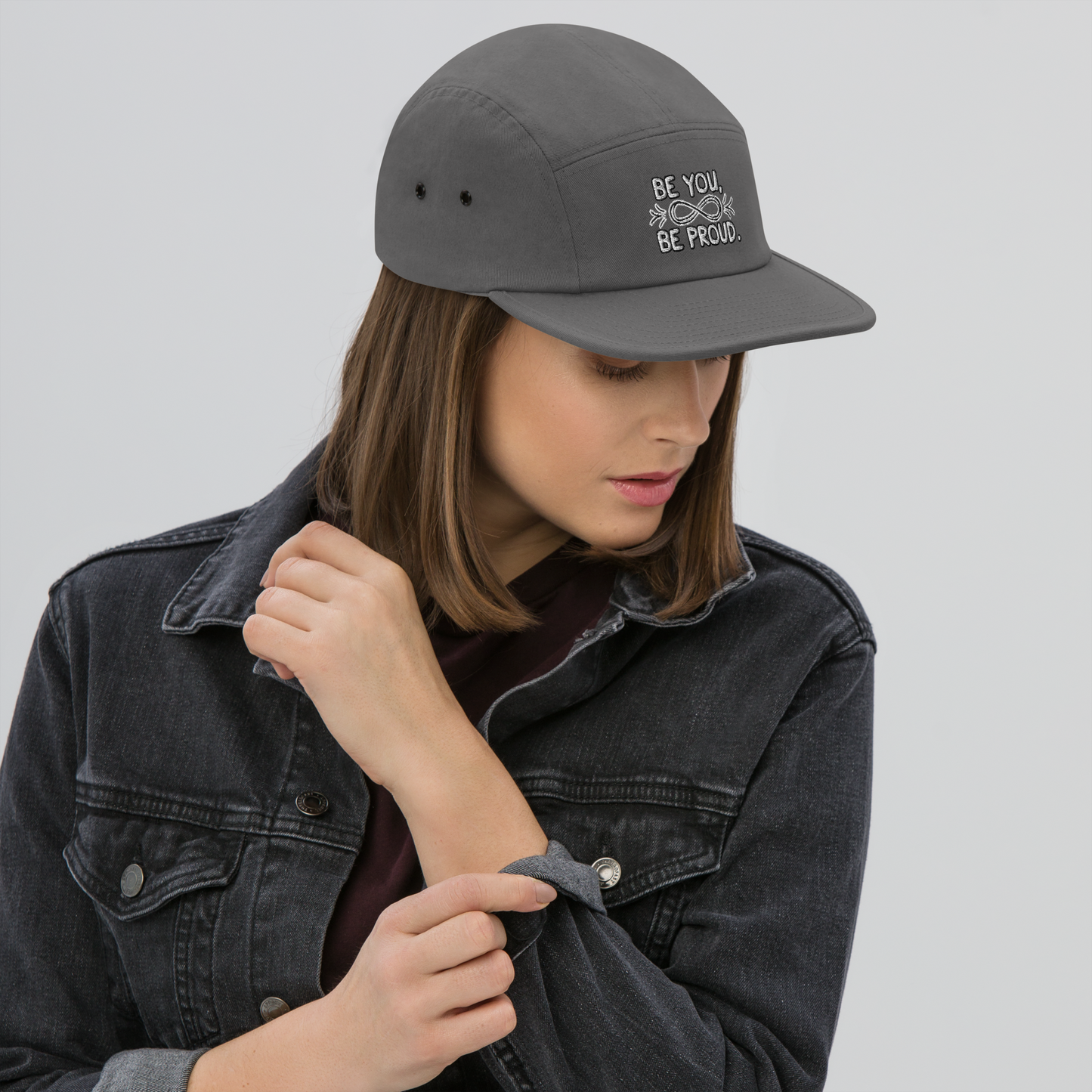 Woman wearing a grey cap with embroidered text 'Be You, Be Proud' and an infinity symbol with arrows, adjusting her denim jacket.