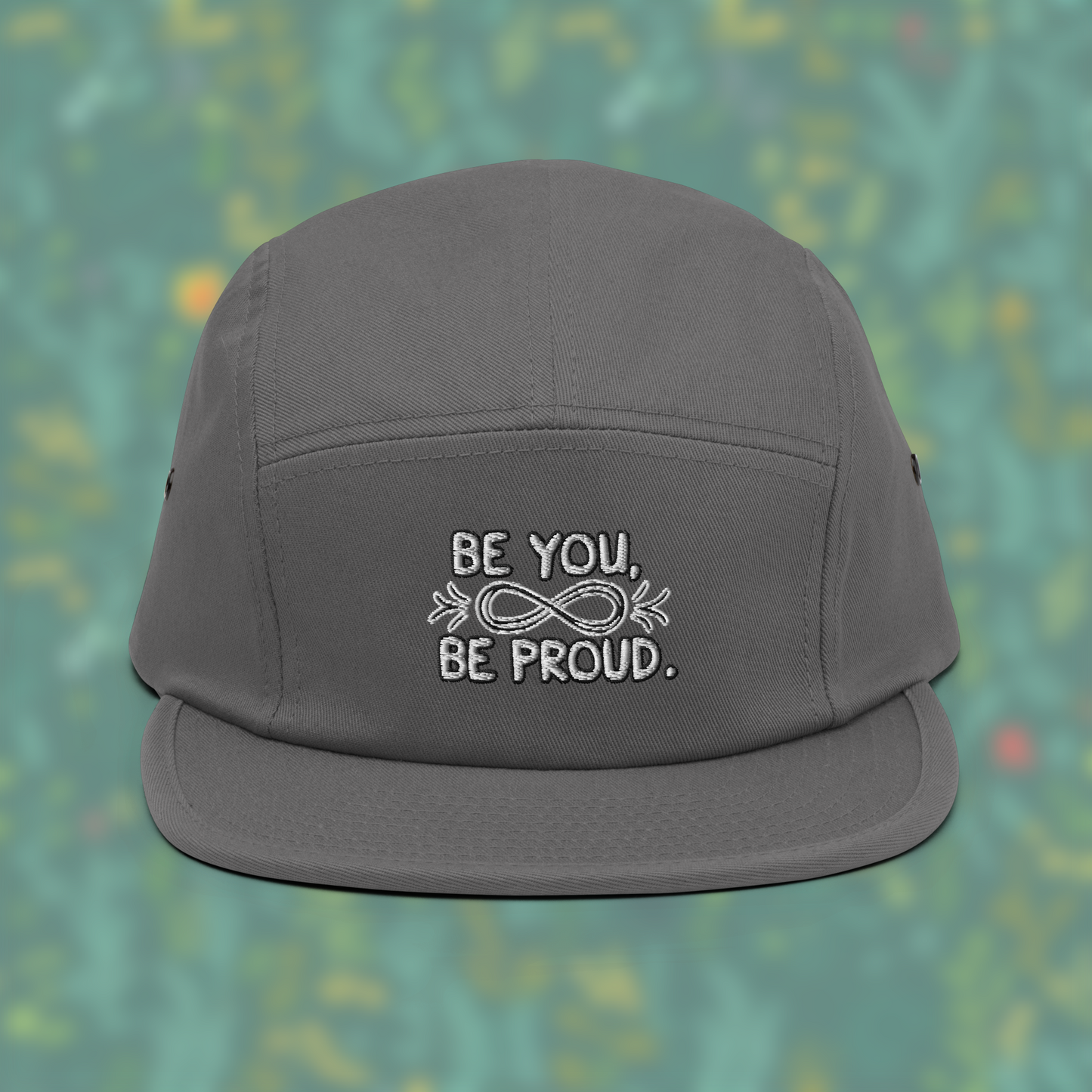 Grey cap with embroidered text 'Be You, Be Proud' and an infinity symbol with arrows, set against a blurred background of green and yellow.
