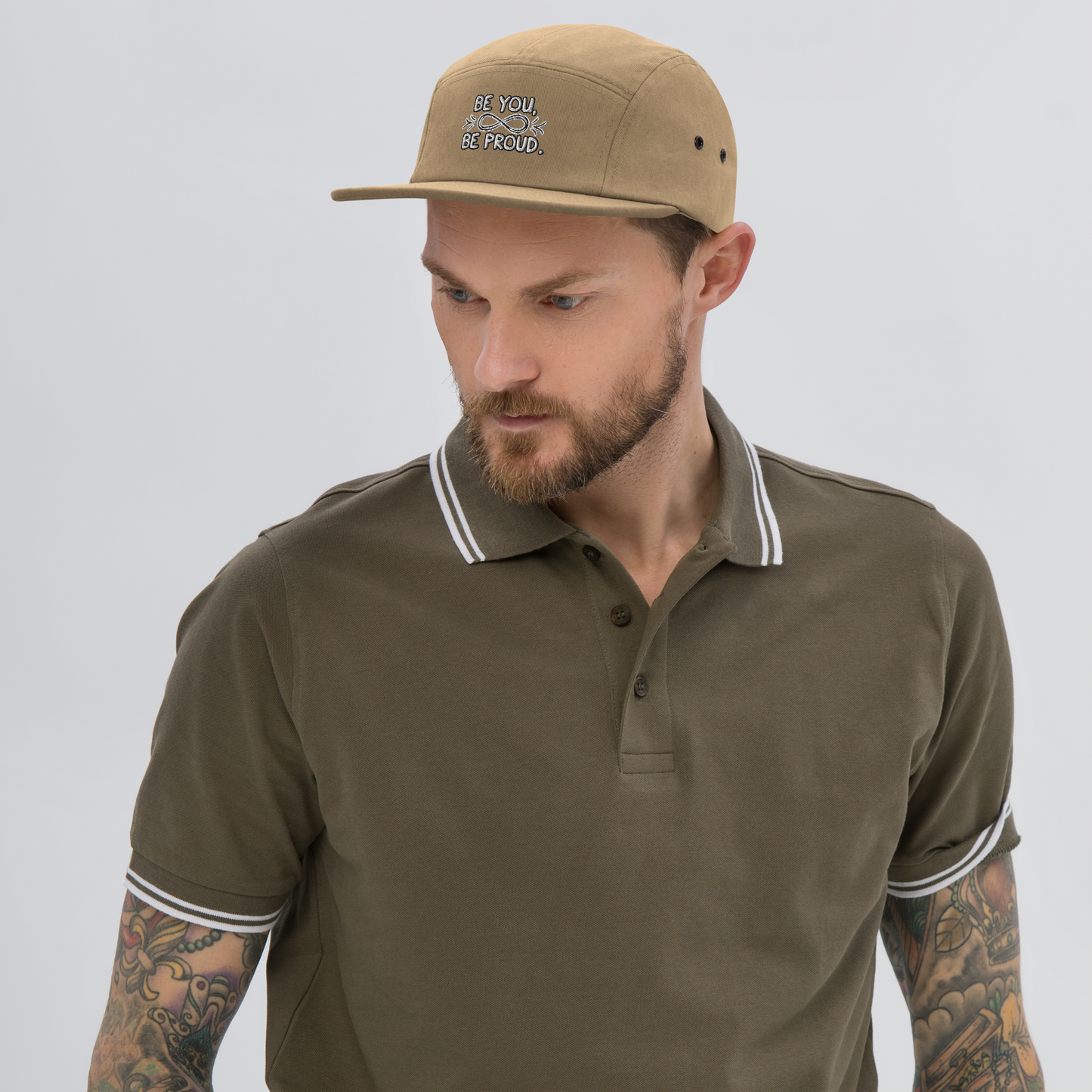 Man wearing a beige cap with embroidered text 'Be You, Be Proud' and an infinity symbol with arrows, looking down while dressed in an olive-green polo shirt.