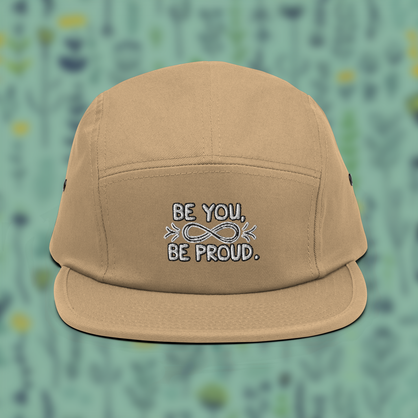 Beige cap with embroidered text 'Be You, Be Proud' and an infinity symbol with arrows, set against a blurred background of green and blue.