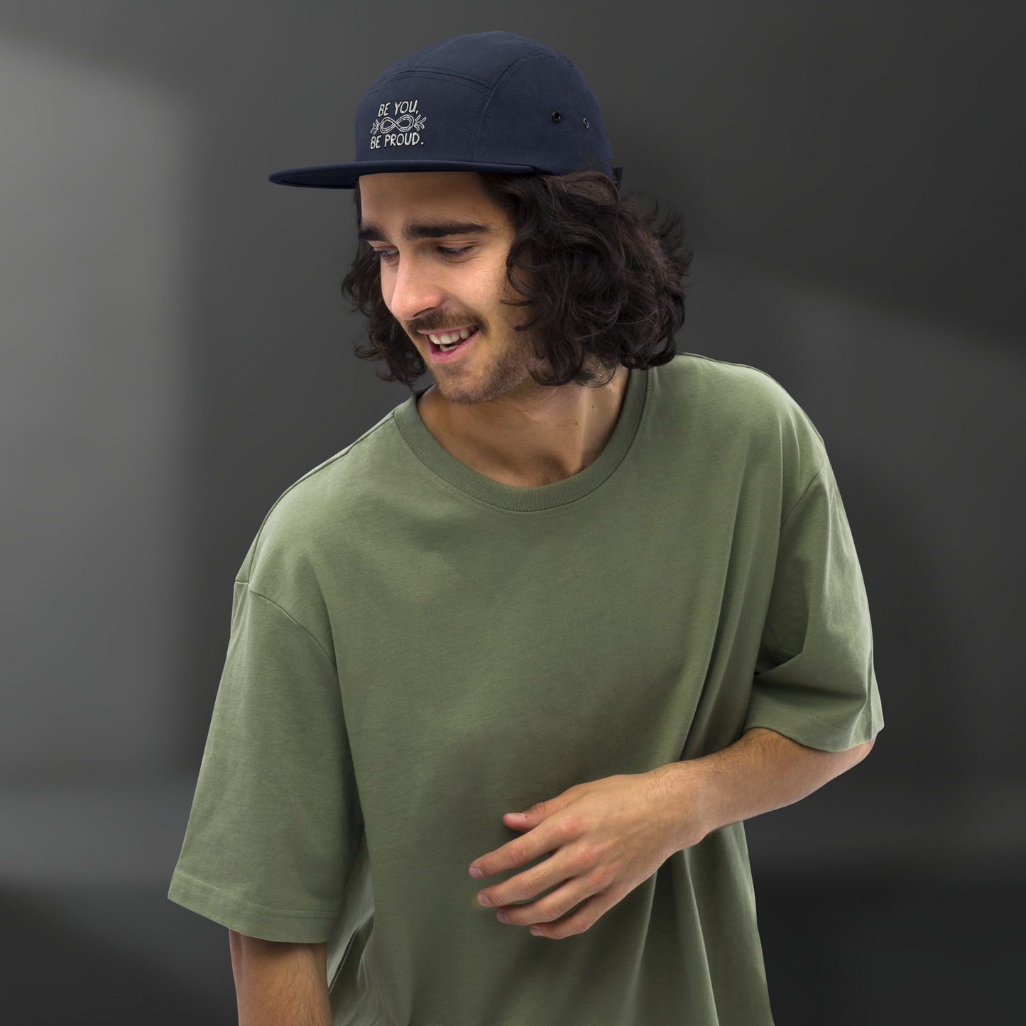 Man wearing a navy blue cap with embroidered text 'Be You, Be Proud' and an infinity symbol with arrows, smiling while wearing an olive-green shirt.