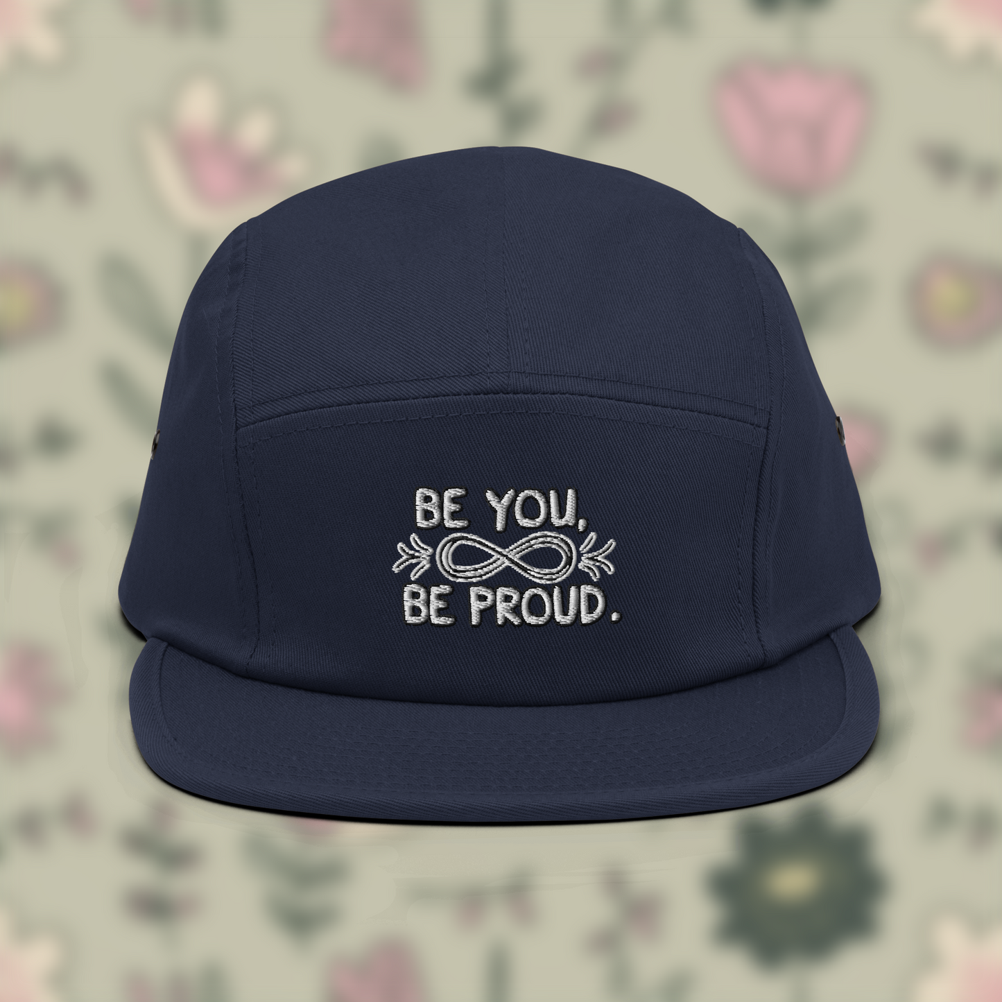 Navy blue cap with embroidered text 'Be You, Be Proud' and an infinity symbol with arrows, set against a blurred background of flowers.