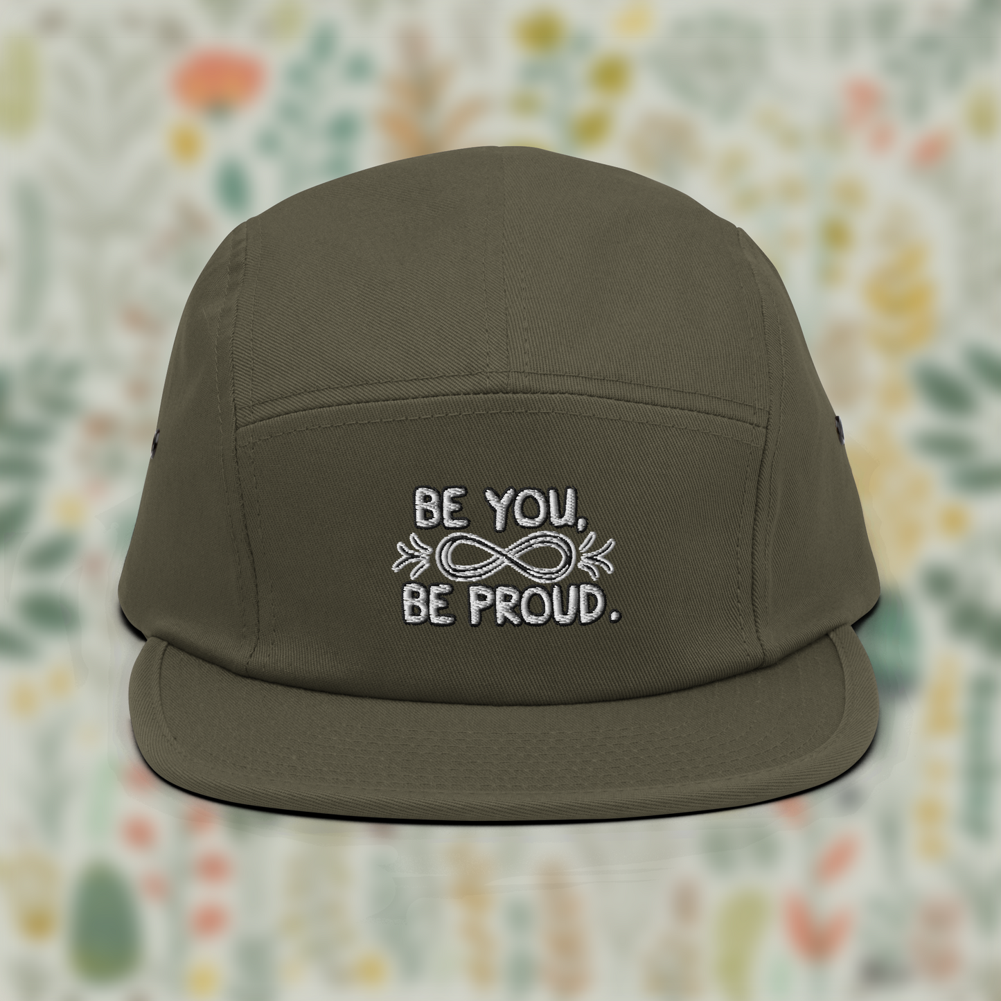 Olive-green cap with embroidered text 'Be You, Be Proud' and an infinity symbol with arrows, set against a blurred background of flowers.