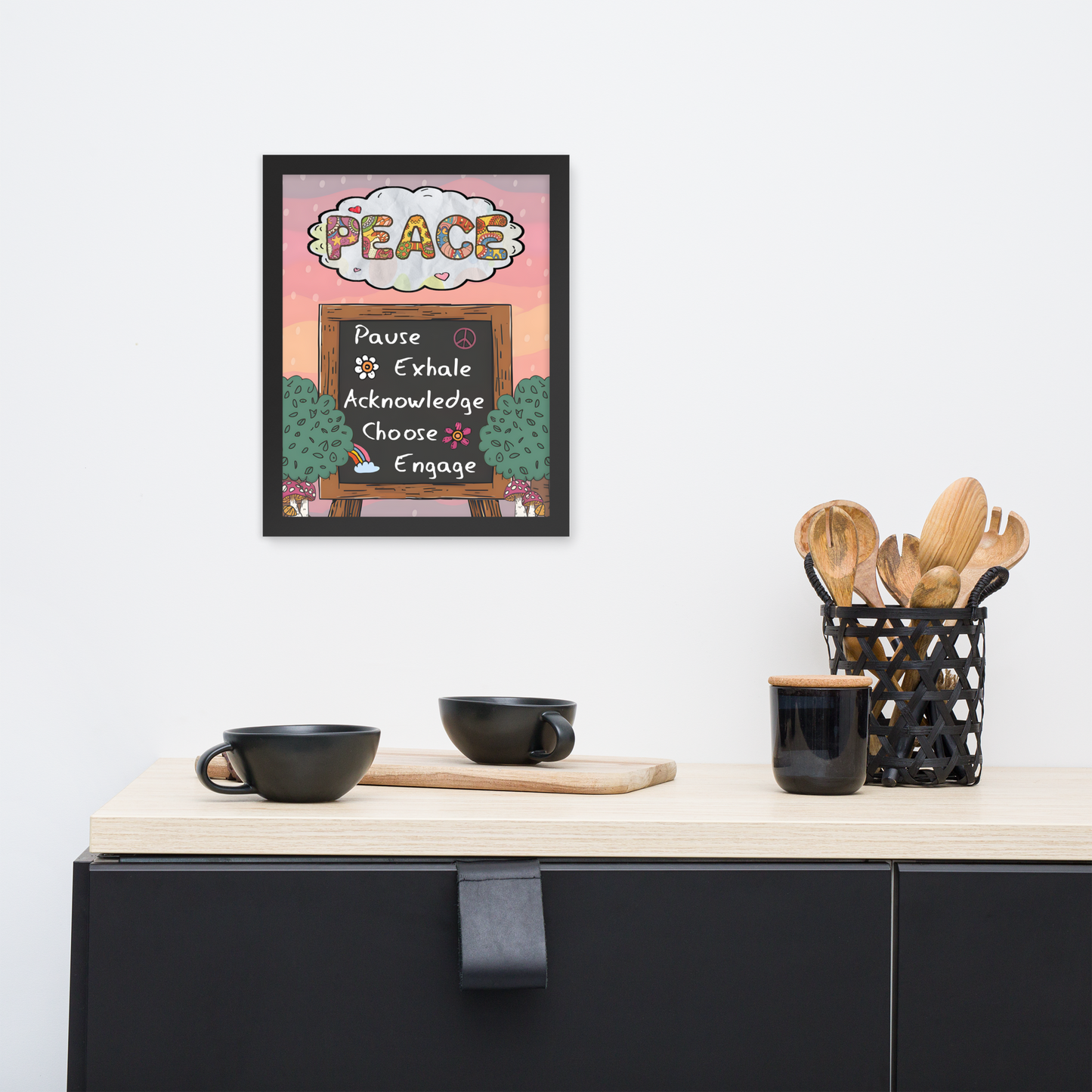 Framed illustrated poster with a black frame titled 'PEACE' featuring a checklist with items: Pause, Exhale, Acknowledge, Choose, Engage. The poster hangs on a white wall above a minimalist kitchen counter with black cups, a wooden cutting board, and a black utensil holder with wooden utensils.