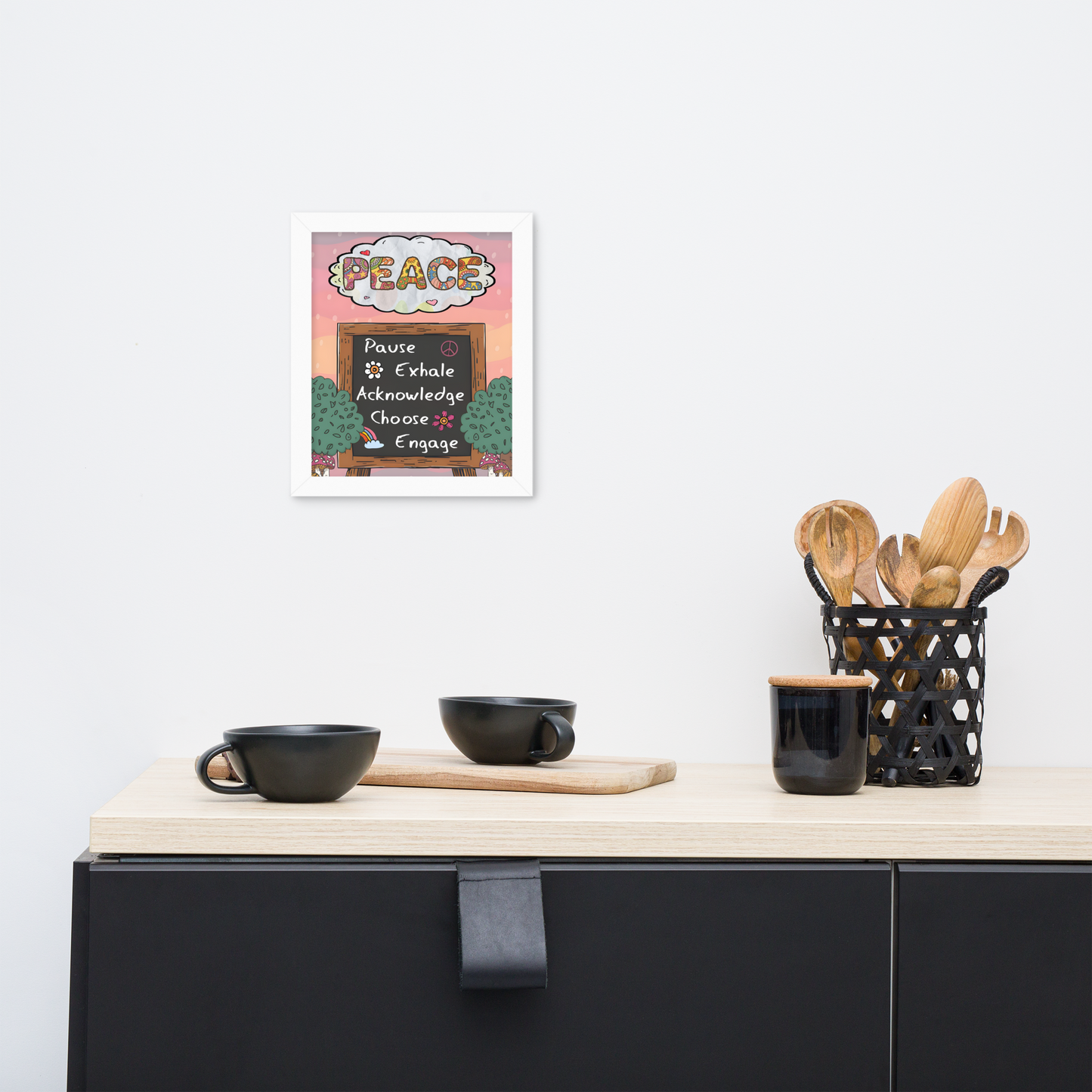 Framed illustrated poster with a white frame titled 'PEACE' featuring a checklist with items: Pause, Exhale, Acknowledge, Choose, Engage. The poster hangs on a white wall above a minimalist kitchen counter with black cups, a wooden cutting board, and a black utensil holder with wooden utensils.