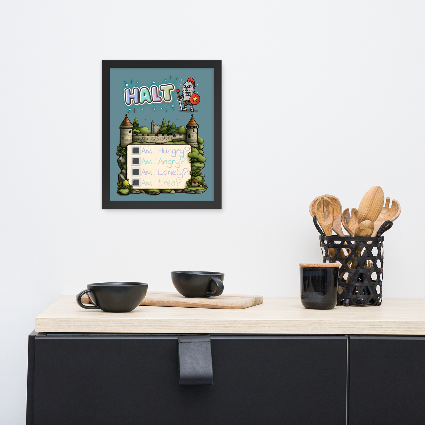 Framed illustrated poster in black frame hanging on a white wall above a minimalist kitchen counter with black cups, a wooden cutting board, and a black utensil holder with wooden utensils. The poster features the word 'HALT' at the top, a cute knight character, a castle background, and a checklist with 'Am I Hungry?', 'Am I Angry?', 'Am I Lonely?', 'Am I Tired?'.