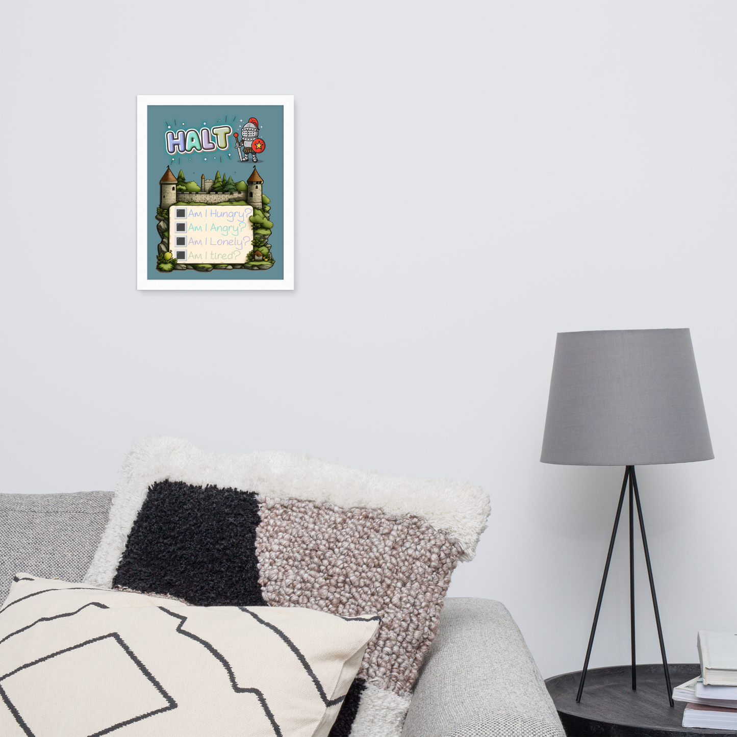 Framed illustrated poster hanging on a white wall above a minimalist sofa and coffee table with a grey lamp. The poster features the word 'HALT' at the top, a cute knight character, a castle background, and a checklist with 'Am I Hungry?', 'Am I Angry?', 'Am I Lonely?', 'Am I Tired?'."