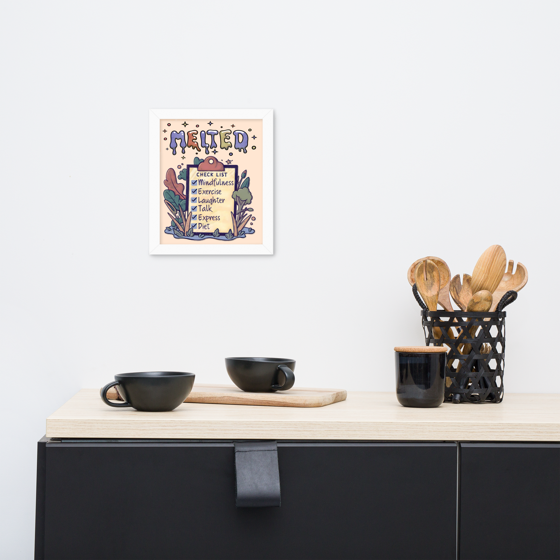 Framed illustrated poster with a white frame titled 'MELTED' featuring a checklist with items: Mindfulness, Exercise, Laughter, Talk, Express, Diet. The poster is hanging on a white wall above a minimalist kitchen counter with black cups, a wooden cutting board, and a black utensil holder with wooden utensils.