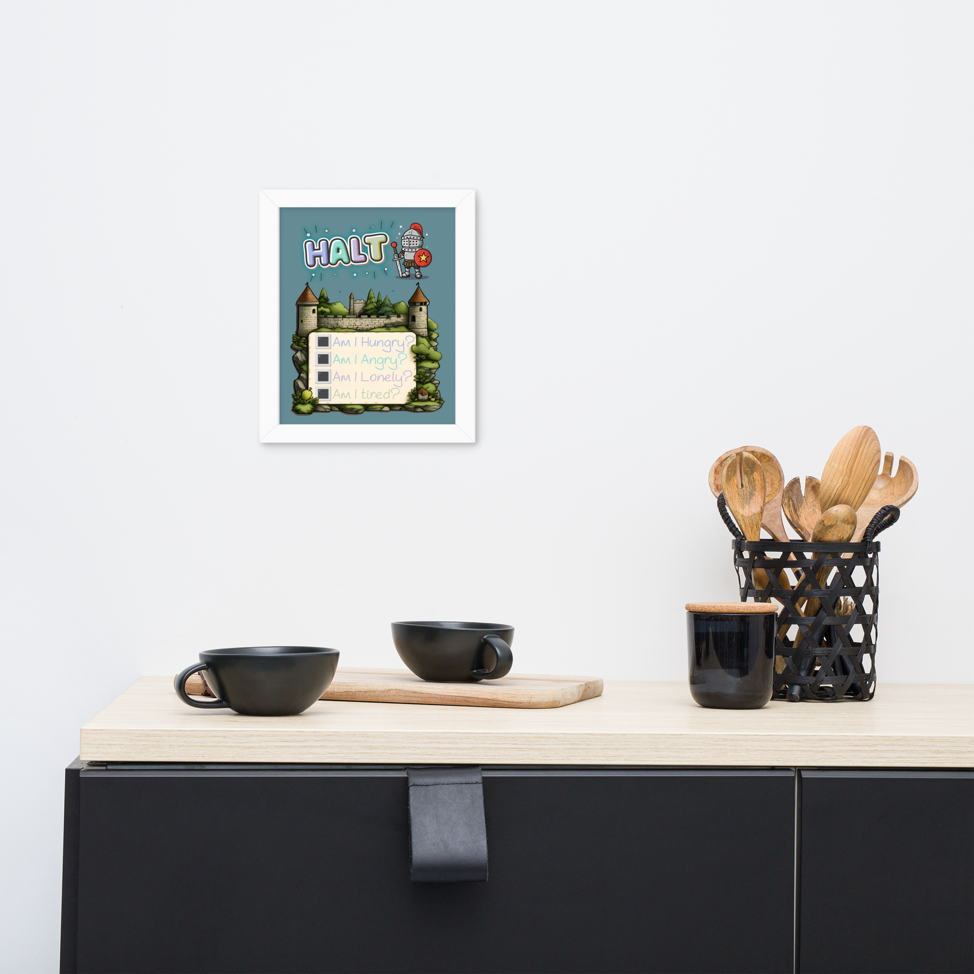 Framed illustrated poster hanging on a white wall above a minimalist kitchen counter with black cups, a wooden cutting board, and a black utensil holder with wooden utensils. The poster features the word 'HALT' at the top, a cute knight character, a castle background, and a checklist with 'Am I Hungry?', 'Am I Angry?', 'Am I Lonely?', 'Am I Tired?'.