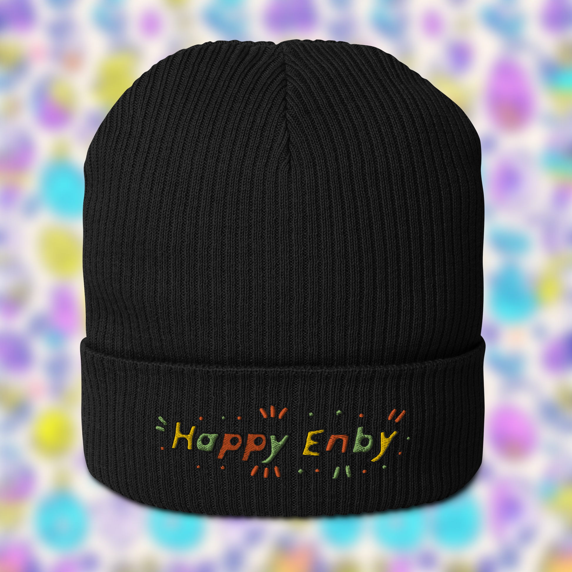 Black beanie with embroidered text 'Happy Enby' in colourful letters, set against a blurred background of pastel colours.