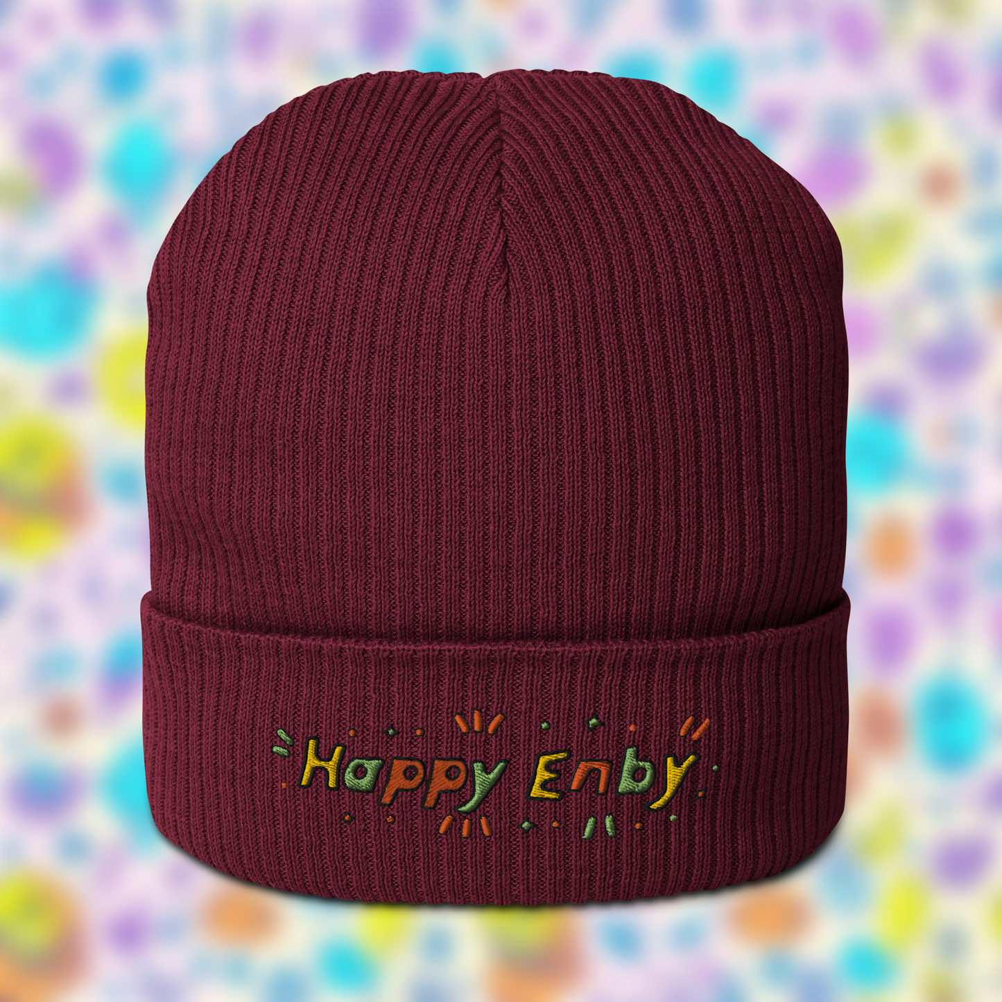 Burgundy beanie with embroidered text 'Happy Enby' in colourful letters, set against a blurred background of pastel colours.