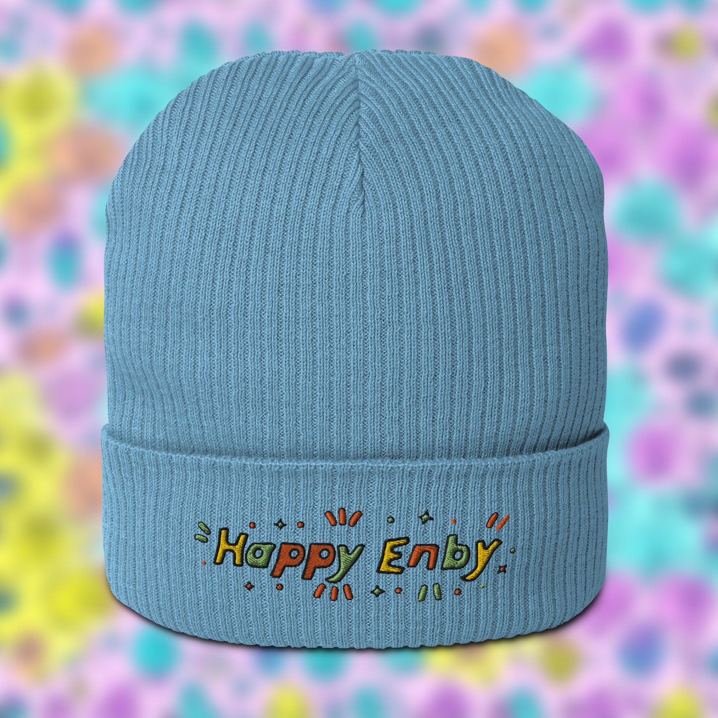 Light blue beanie with embroidered text 'Happy Enby' in colourful letters, set against a blurred background of pastel colours.