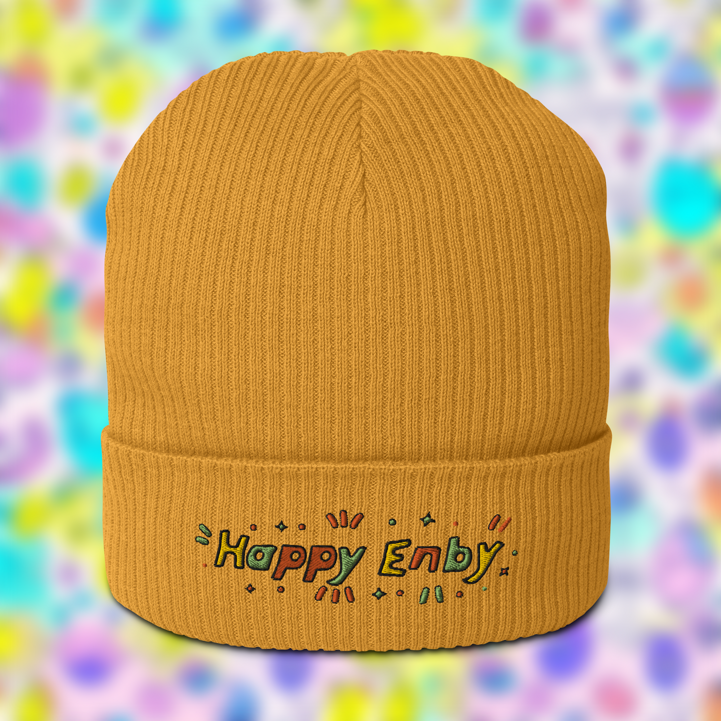 Yellow beanie with embroidered text 'Happy Enby' in colourful letters, set against a blurred background of pastel colours.